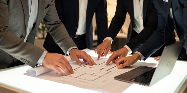 Un couple examine des plans et des documents immobiliers sur une table lumineuse, entouré de plusieurs professionnels dans un bureau moderne.