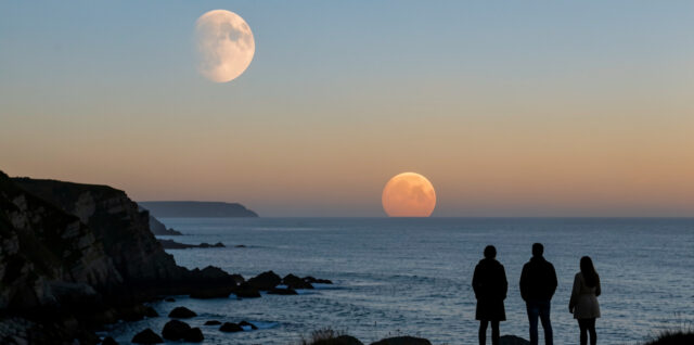 Éclipse lunaire partielle, Lune cuivrée basse à l’ouest à l’aube au-dessus d’un littoral français, avec trois observateurs en silhouette et une mer calme.