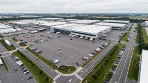 Vue aérienne réaliste d’un grand hypermarché français avec parking, bâtiment commercial, accès routiers et zones logistiques dans une ambiance sobre.